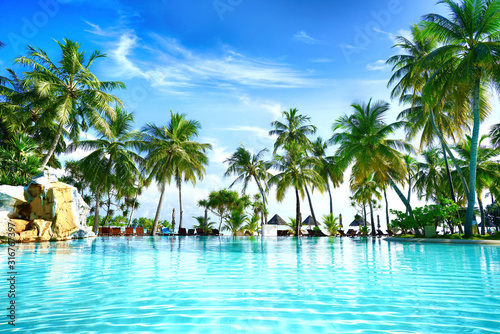 Beautiful lush tropical palm trees against blue sky with white clouds are reflected in turquoise textured wavy water on sunny day. Colorful image for summer vacation.
