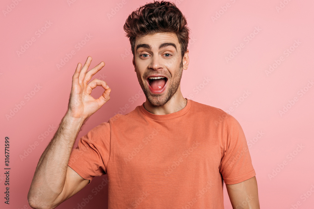 cheerful young man showing okay gesture while looking at camera on pink background cheerful young man showing okay gesture while looking at camera on pink background