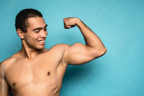 Beauty portrait of half naked handsome young man with beautiful torso dressed in towel touching his biceps looking at camera isolated over blue background