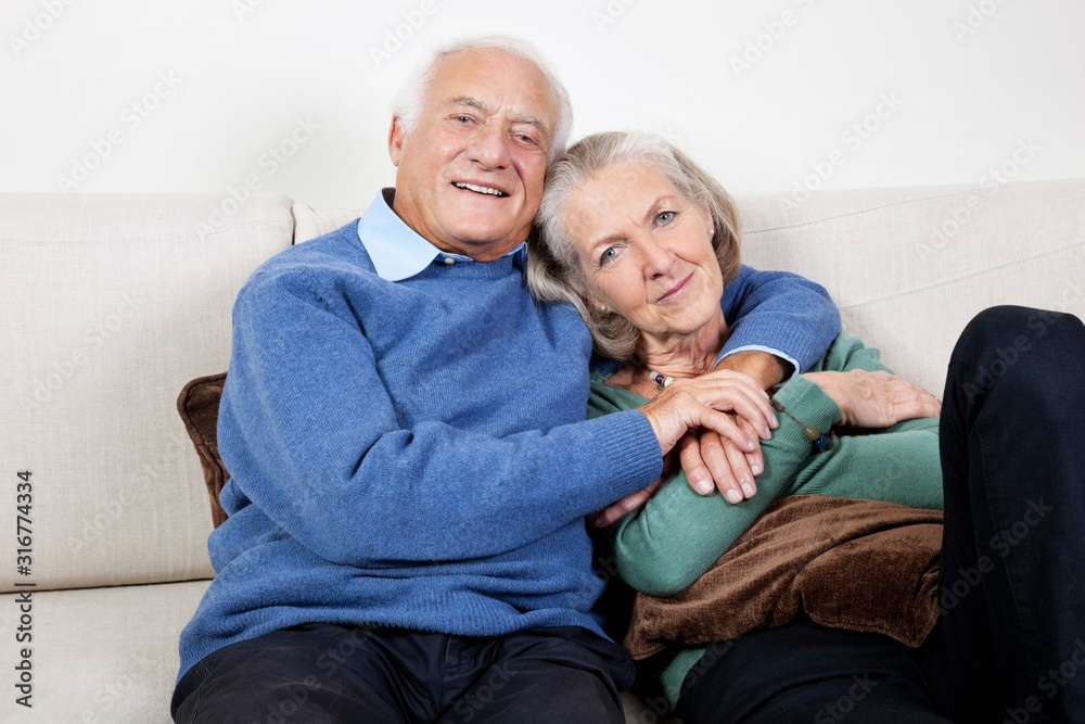 Portrait of happy elderly man with arm around spouse sitting on sofa at home