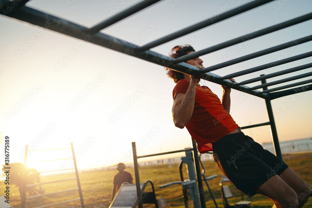 Fit young man doing pull-ups at an outdoor gym park during bright hot summers day