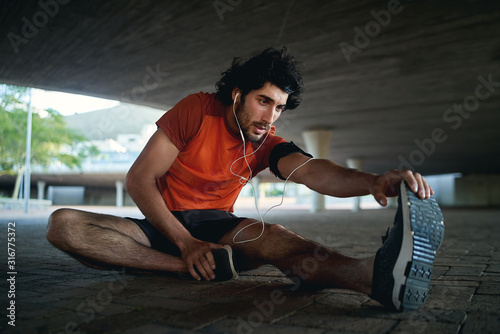 Young man athlete with earphone in his ears warming up and stretching his legs before running in the city street under the bridge