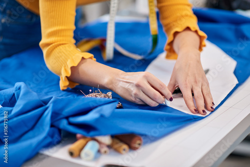 Close up of creative caucasian fashion designer standing in her studio and drawing scheme on blue linen for a beautiful evening dress.