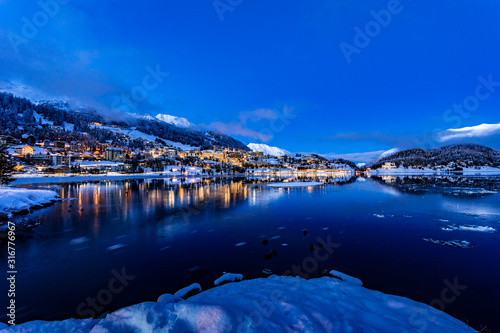 View of beautiful night lights of St. Moritz town in Switzerland at night in winter, with reflection from the lake and snow mountains in backgrouind