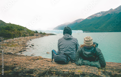 couple of tourists resting sitting on a rock on the shore of a blue lake surrounded by mountains and trees with the Perito Moreno glacier in the background under a cloudy sky