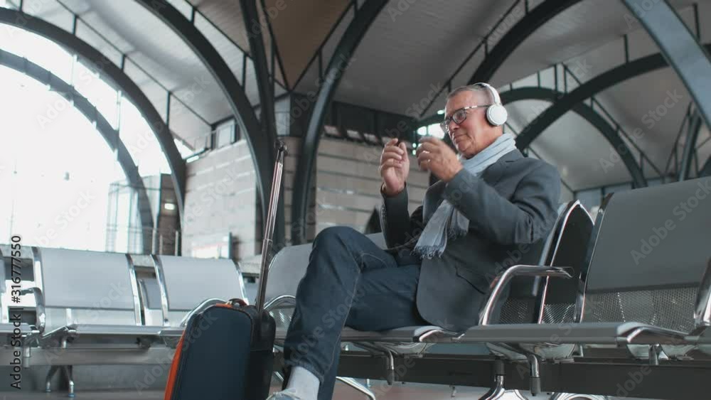 Senior tourist in the airport waits for his flight and listens to music with headphones Senior tourist in the airport waits for his flight and listens to music with headphones