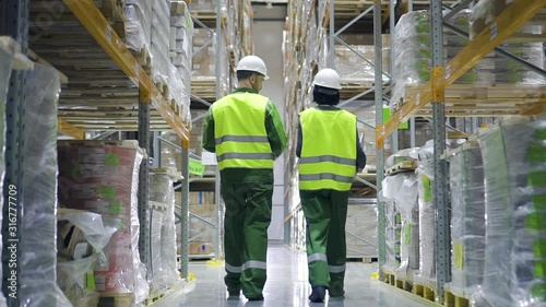 Young people workers talking and walking on industrial warehouse during working day. Back view of american man and woman having conversation about work in plant storehouse with products on shelves