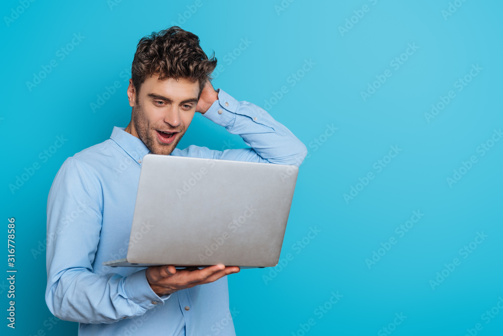 surprised young man touching head while looking at laptop on blue background - obrazy, fototapety, plakaty 