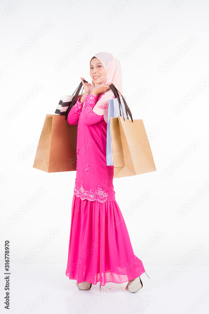 A beautiful and excited Muslim female model in a traditional pink modern kurung carrying shopping bags isolated on white background. Eidul fitri fashion and festive shopping concept