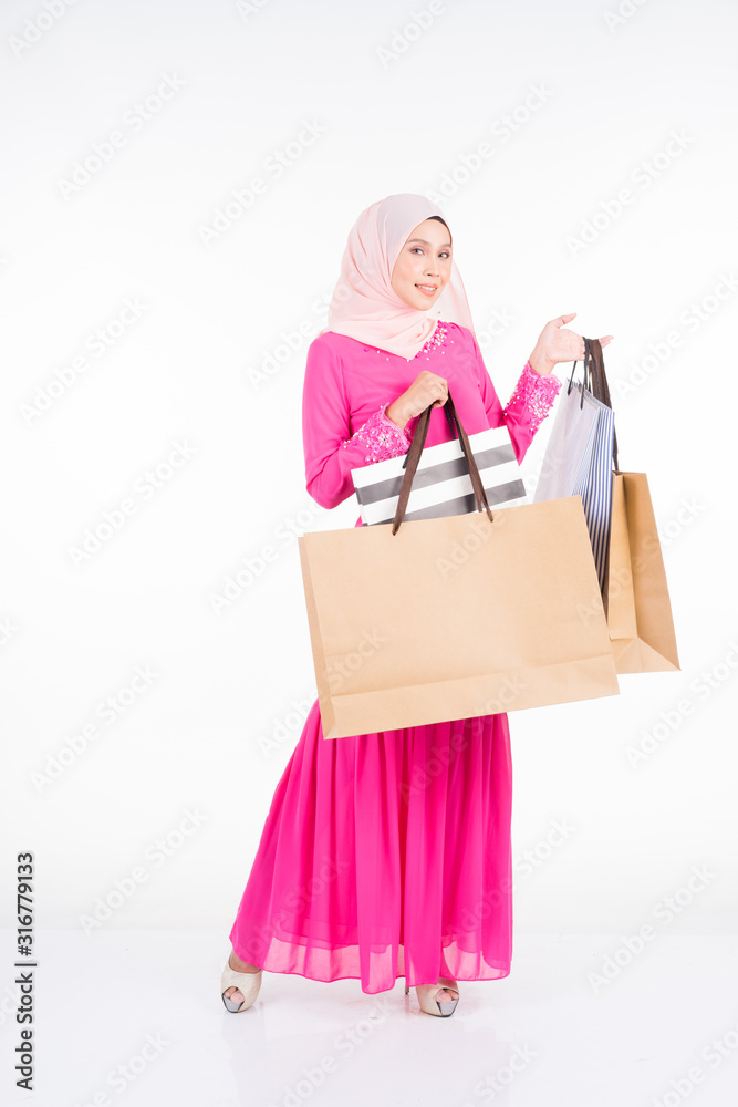 A beautiful and excited Muslim female model in a traditional pink modern kurung carrying shopping bags isolated on white background. Eidul fitri fashion and festive shopping concept A beautiful and excited Muslim female model in a traditional pink modern kurung carrying shopping bags isolated on white background. Eidul fitri fashion and festive shopping concept