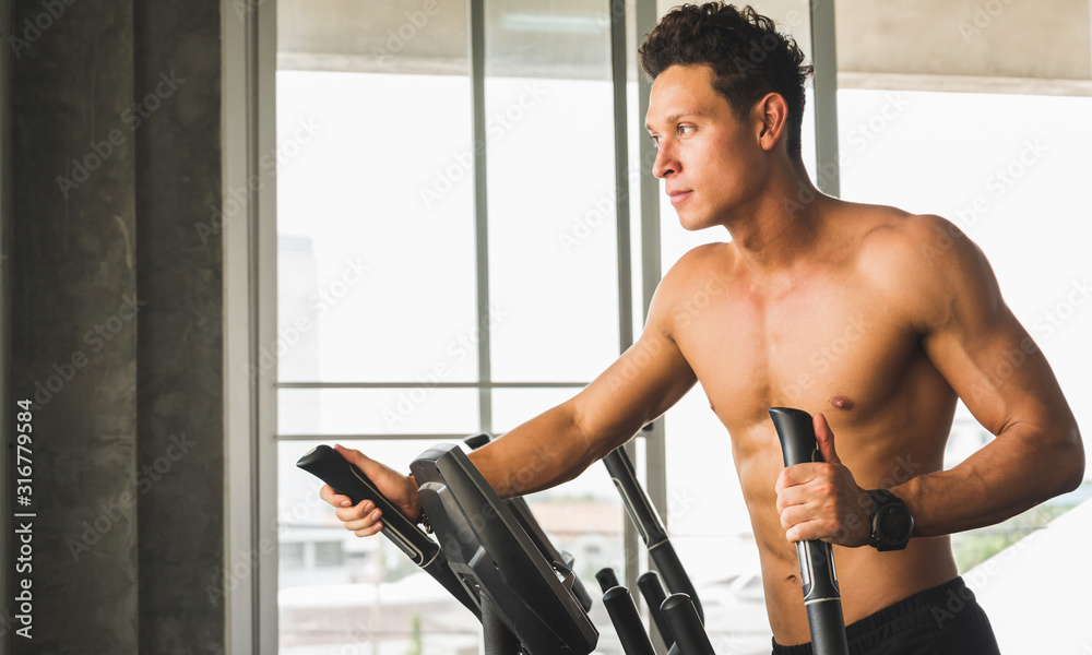 Obraz premium Young man at the gym exercising on the cross trainer machine. Fitness man doing cardio workout program.