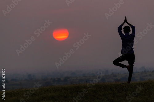 Silhouette of a man doing yoga in front of the sun during the sunrise with one leg raised and arms above the head praying