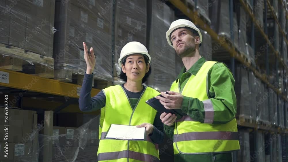 Young american employees having conversation, standing in warehouse during working day. Front view of man and woman talking about work on background of shelving with boxes, male worker holding tablet Young american employees having conversation, standing in warehouse during working day. Front view of man and woman talking about work on background of shelving with boxes, male worker holding tablet