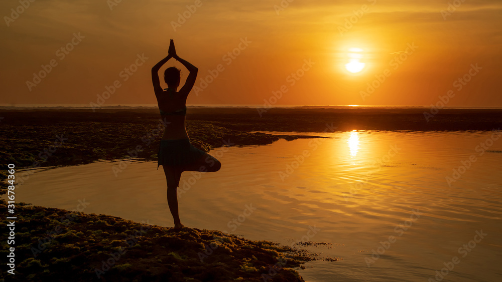 Vrikshasana asana. Young woman practicing tree pose at the beach during sunset. Arms raising with namaste mudra. Melasti beach, Bali. View from back.
