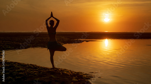 Vrikshasana asana. Young woman practicing tree pose at the beach during sunset. Arms raising with namaste mudra. Melasti beach, Bali. View from back.