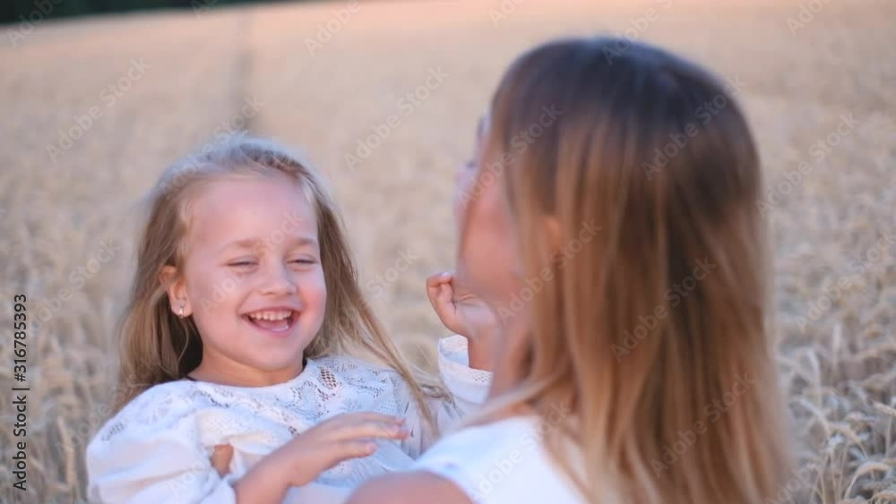 A young mother holds her little daughter in her arms and hugs her in a wheat field in summer at sunset. A young mother holds her little daughter in her arms and hugs her in a wheat field in summer at sunset.