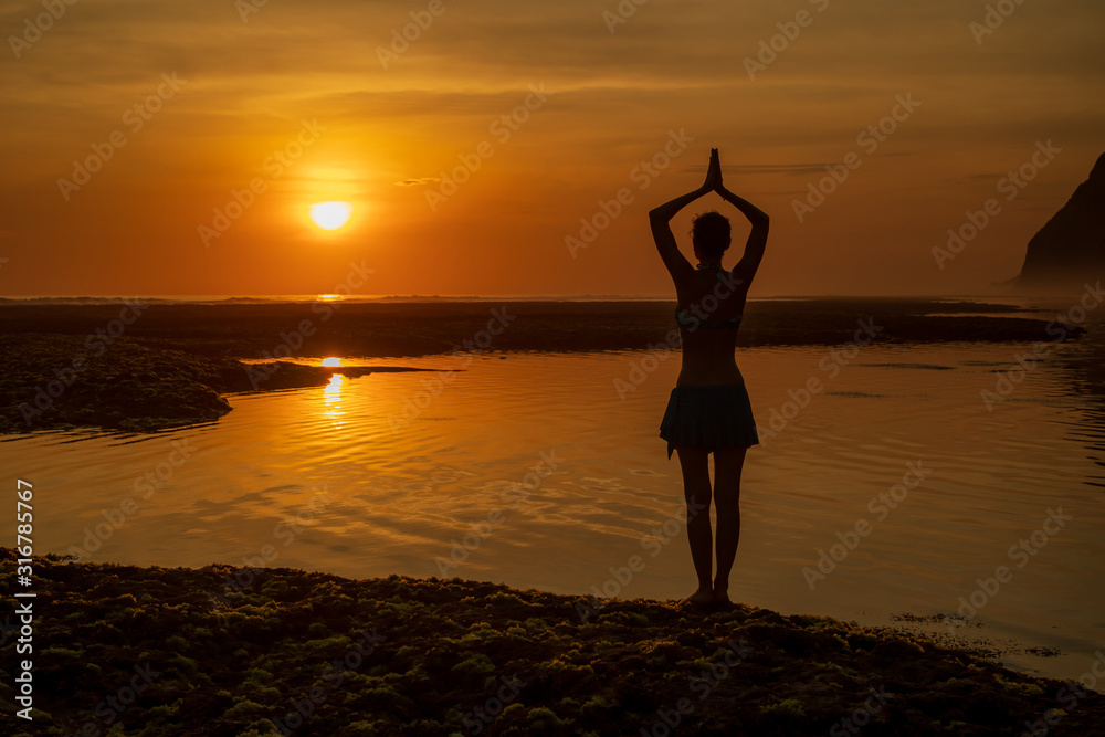Yoga pose. Woman standing on the beach, practicing yoga. Young woman raising arms with namaste mudra during sunset golden hour. View from back. Melasti beach, Bali.