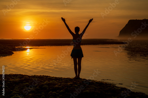 Excited young woman raising arms at the beach in front of the ocean. View from back. Sunset golden hour at the beach. Bali, Indonesia.