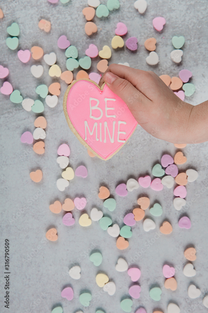 Fototapeta premium Valentine's Day hand holding a be Mine Heart Shaped pink sugar cookie and scattered conversation hearts scattered on a gray cement neutral background