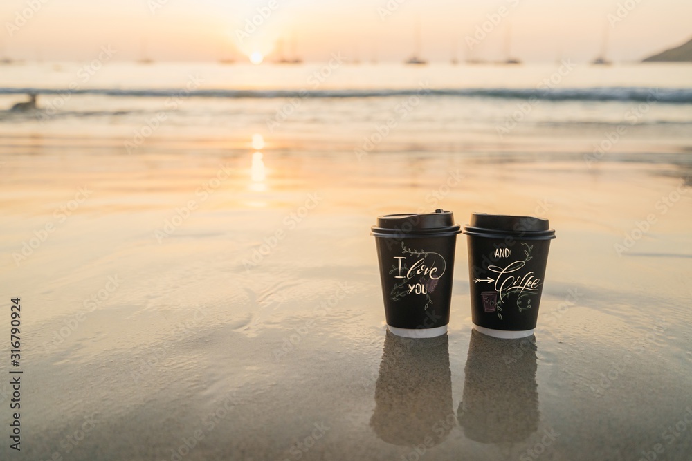 two cups of disposable black paper stand on the sand by the sea during sunset. Romantic concept for valentines day. two cups of disposable black paper stand on the sand by the sea during sunset. Romantic concept for valentines day.