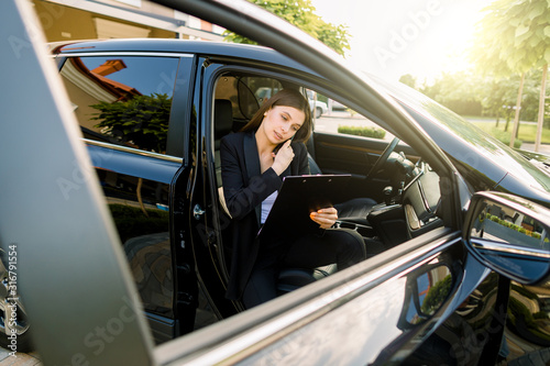 Side angle view of young pretty businesswoman, sitting inside her car on passenger seat, while using a digital tablet and talking mobile phone