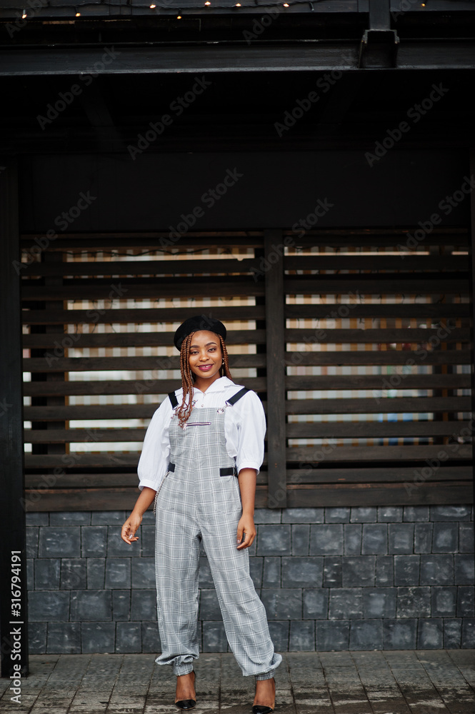Fototapeta premium African american woman in overalls and beret posed outdoor against wooden background.