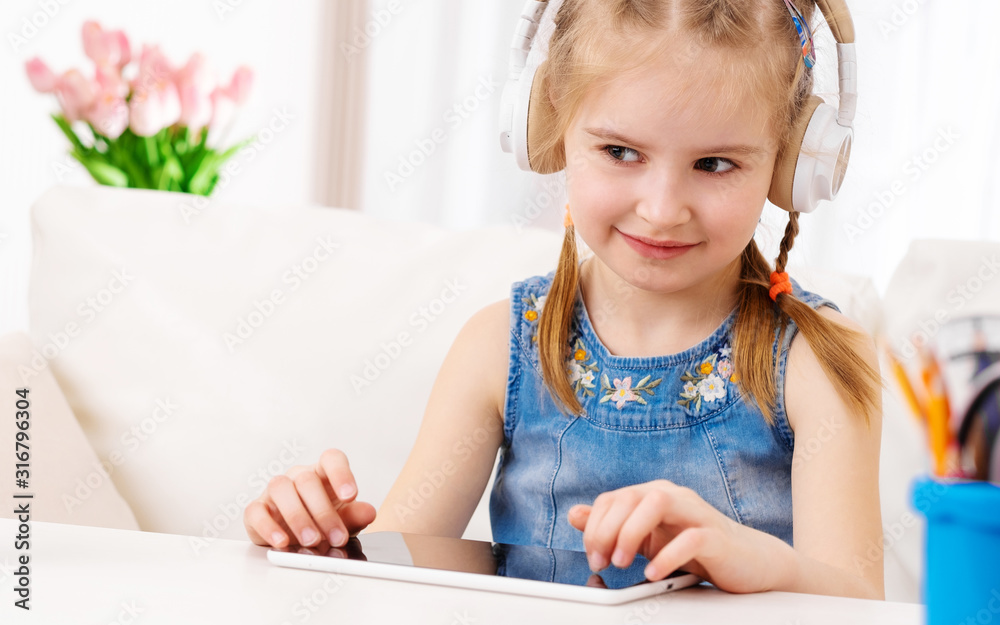 Beatiful little girl with white tablet with headphones on, in the room Beatiful little girl with white tablet with headphones on, in the room