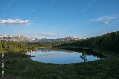 Clouds and moon reflected in a mountain lake. A calm summer evening. A chain of snow-capped mountains. Altai.