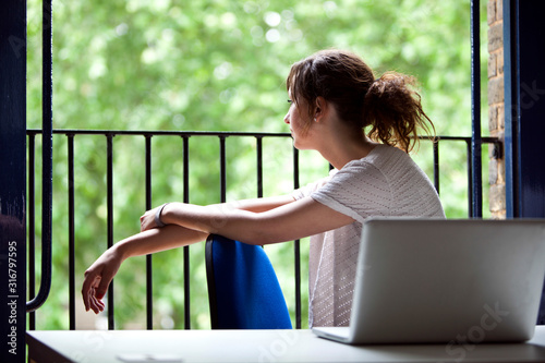Thoughtful young businesswoman sitting at desk by balcony