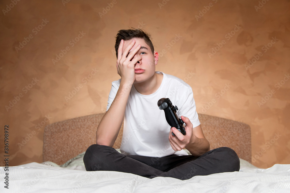 Fototapeta premium The teenager is upset, making face palm, in his second hand he has a joystick