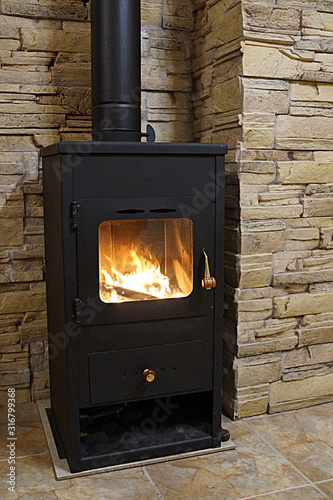  metal stove fireplace with fire behind glass in the interior against the background of a wall of natural stone