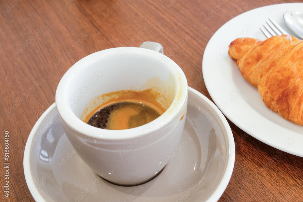 Selective focus of white ceramic cup of espresso coffee shot place on nice and clean wooden table with fresh baked croissant blurred as background. Morning breakfast for refreshment or diet food. Selective focus of white ceramic cup of espresso coffee shot place on nice and clean wooden table with fresh baked croissant blurred as background. Morning breakfast for refreshment or diet food.