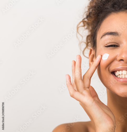 Fotografia Face care. Happy black woman applying moisturizer cream on cheek