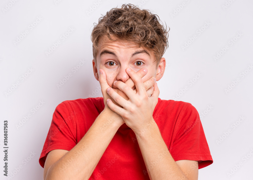Portrait of scared teen boy on grey background. Funny child looking at camera in shock or amazement covering mouth with hands. Handsome caucasian young teenager posing at studio. Portrait of scared teen boy on grey background. Funny child looking at camera in shock or amazement covering mouth with hands. Handsome caucasian young teenager posing at studio.