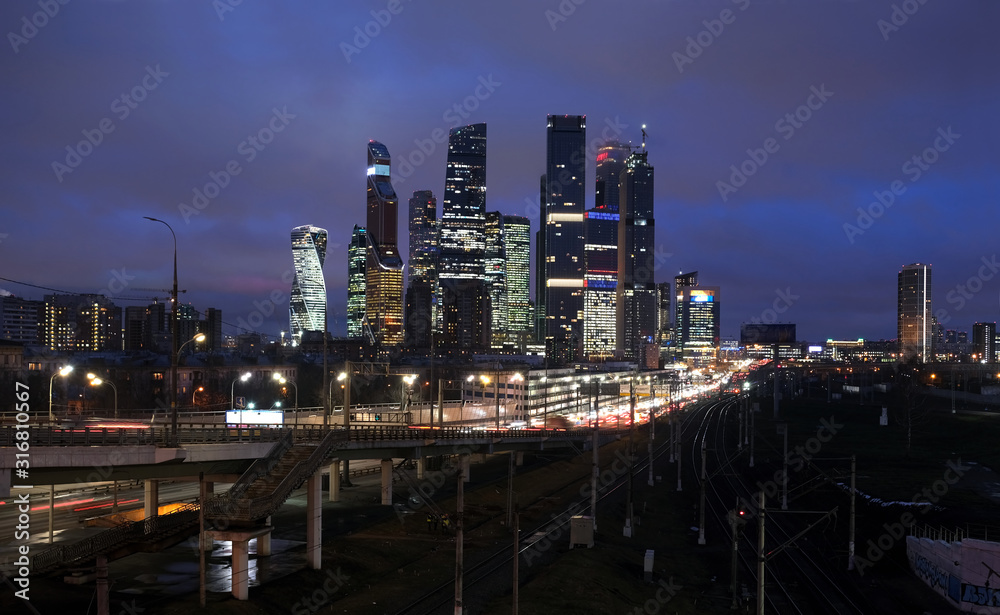 City landscape with many railroad tracks going far, cars on the overpass and skyscrapers with towers of Moscow CIty with lighting windows in the evening