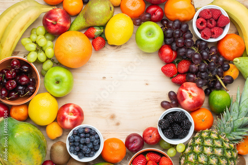 Rainbow fruits background circle frame  strawberries raspberries oranges plums apples kiwis grapes blueberries mango persimmon on light wooden table  top view  copy space for text  selective focus