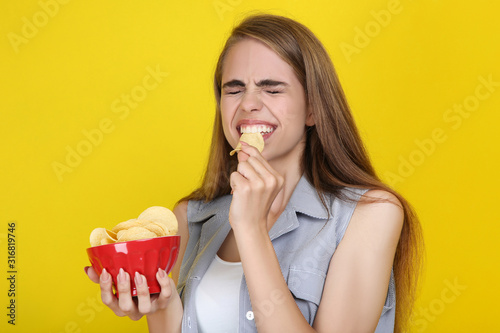 Young beautiful girl eating potato chips on yellow background