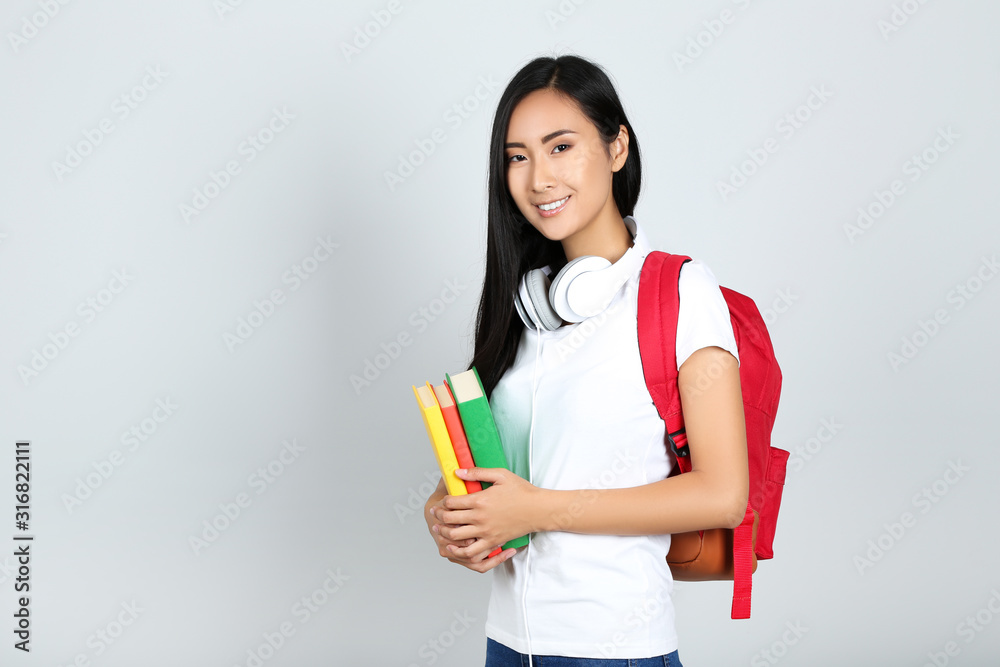 Young woman with books, headphones and backpack on grey background