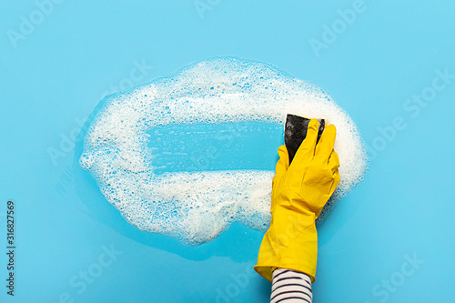 Foto Hand in a yellow rubber glove holds a cleaning sponge and wipes a soapy foam on a blue background