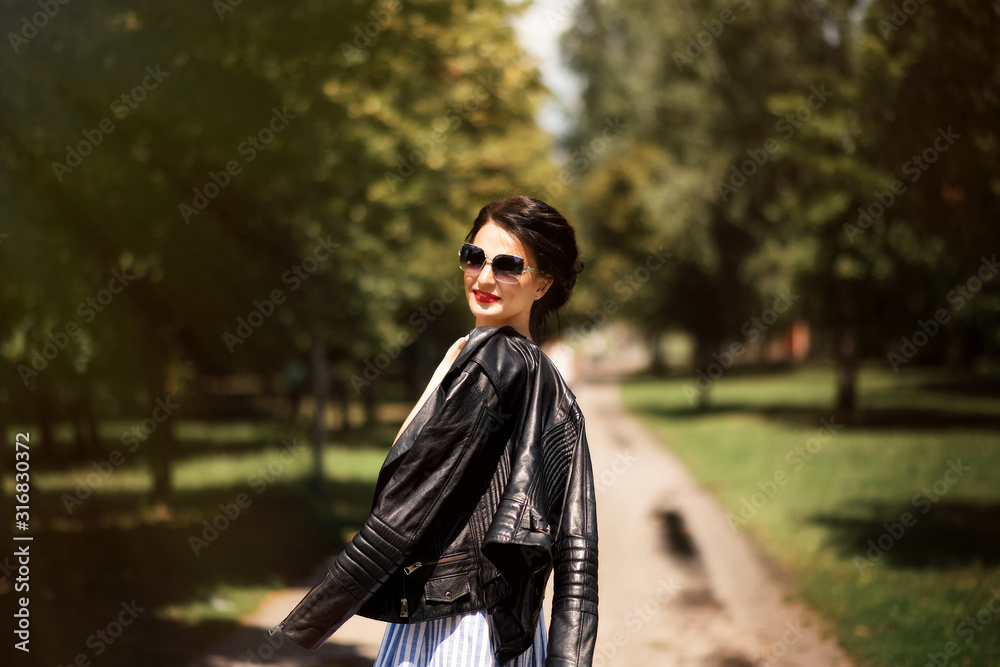 Portrait of beautiful stylish brunette girl in blue and white striped sleeveless dress and sunglasses with black leather jacket in hands in park in summer time. Summer fashion lifestyle.