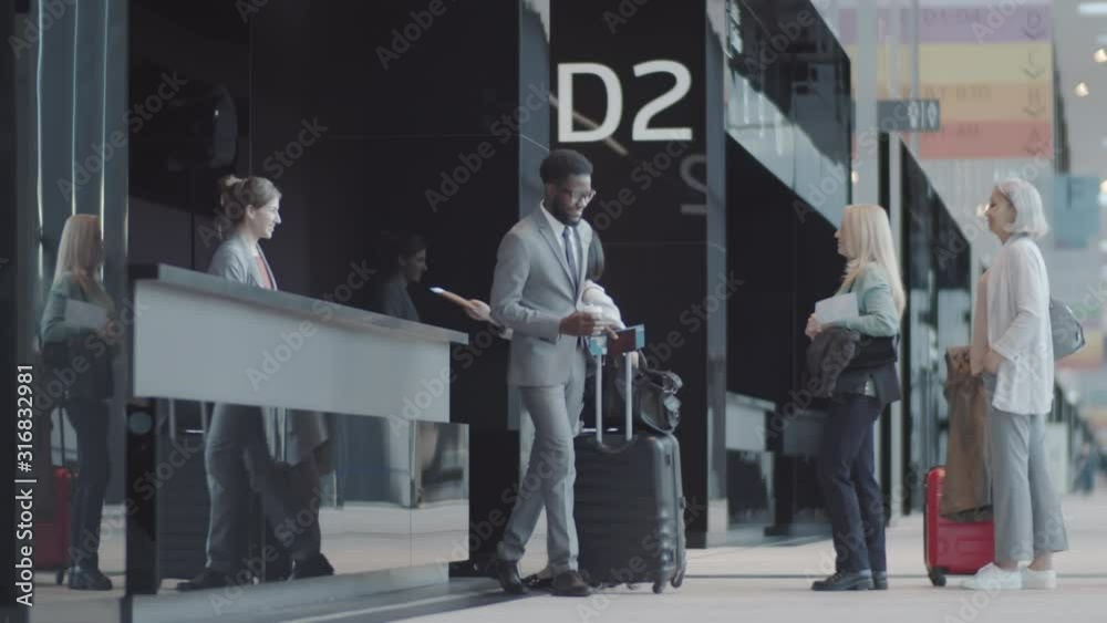 Wide shot of diverse passengers with suitcases standing in queue at check-in counter at airport, giving passport and ticket to smiling female agent, talking, thanking and walking away