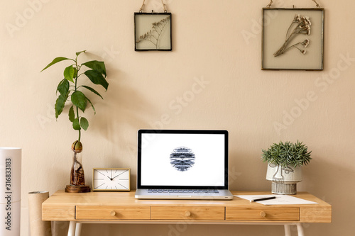Stylish and creative wooden desk with laptop mock up screen, avocado plant office accessories, plant and gold clock. Beige background wall. Design home office interior. Template.
