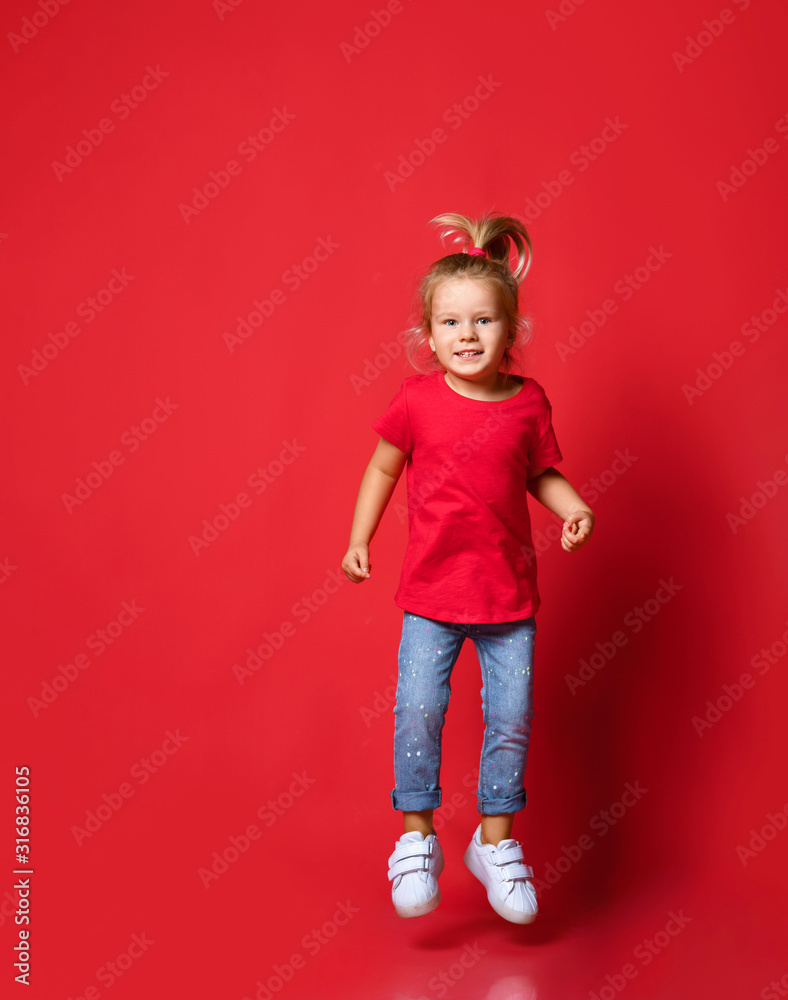 Small happy excited blond girl in stylish casual clothing and white sneakers jumping over red wall background Small happy excited blond girl in stylish casual clothing and white sneakers jumping over red wall background