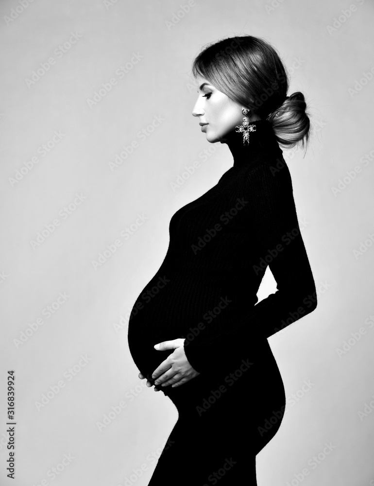 Young beautiful pregnant woman in black dress and massive earrings standing and looking down  - obrazy, fototapety, plakaty 