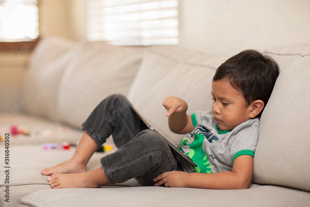Relaxing on a couch at home, a toddler uses his finger to interact with a tablet he is using. Relaxing on a couch at home, a toddler uses his finger to interact with a tablet he is using.