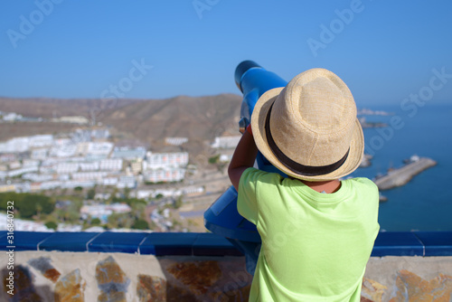 European boy  in sunhat observing beautiful landscape in a spyglass during his summer vacations in Spain.