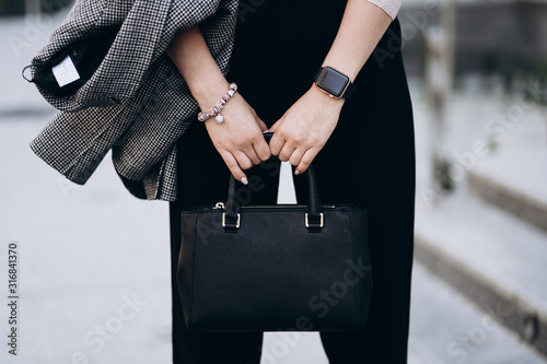 Elegant outfit. Close up of textured big dark bag. Model posing in street, wearing short trousers, creamy sweater, gray coat or jacket and white sneakers . Female fashion concept.