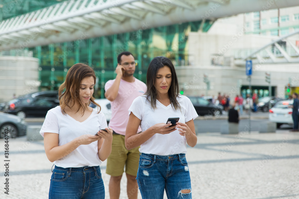 Two women walking on street with phones. Smiling friends holding smartphones while strolling. Communication and technology concept Two women walking on street with phones. Smiling friends holding smartphones while strolling. Communication and technology concept