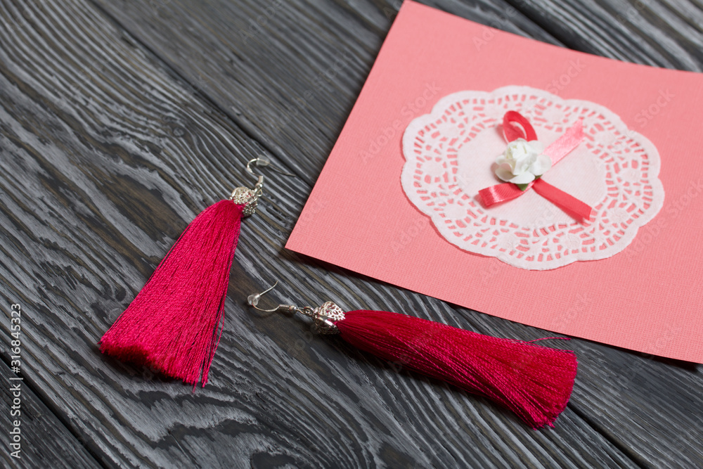 Homemade red tassel earrings and a greeting card. They lie on brushed pine boards painted in black and white.