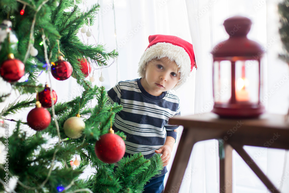 Beautiful blonde toddler boy, decorating christmas tree with balls and light strings
